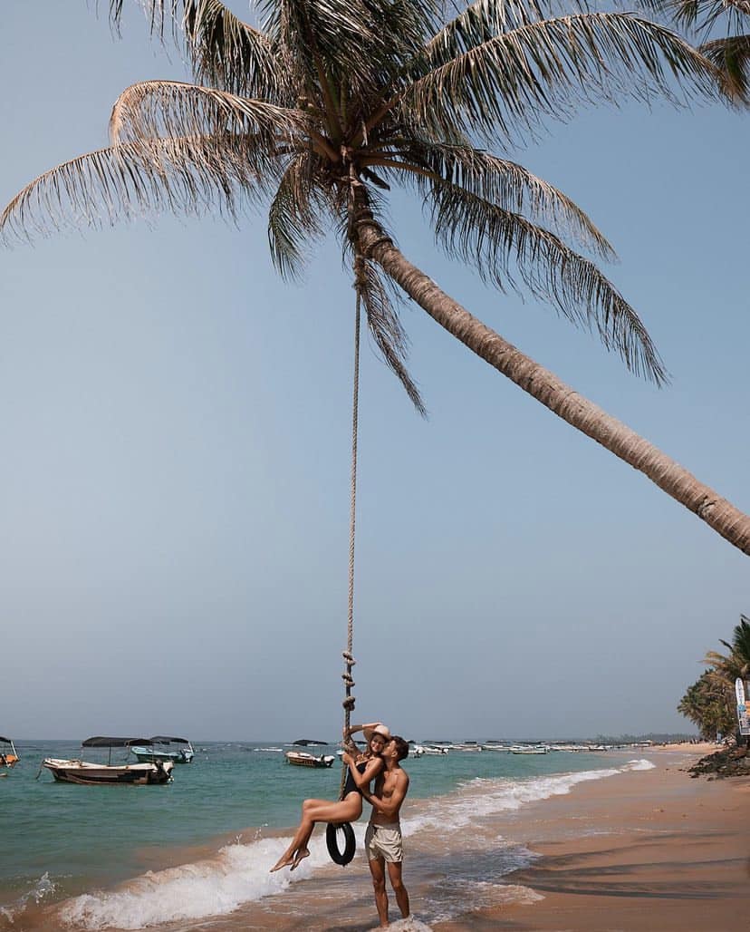 A girl sits on a swing strung to a coconut palm overhanging the beach with her partner standing alongside.