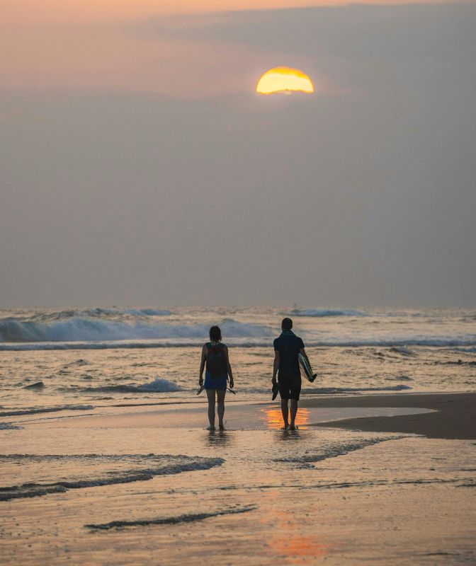 A couple walks towards the tropical sunset on a cloudy afternoon, flip-flops and surfboard in hand.