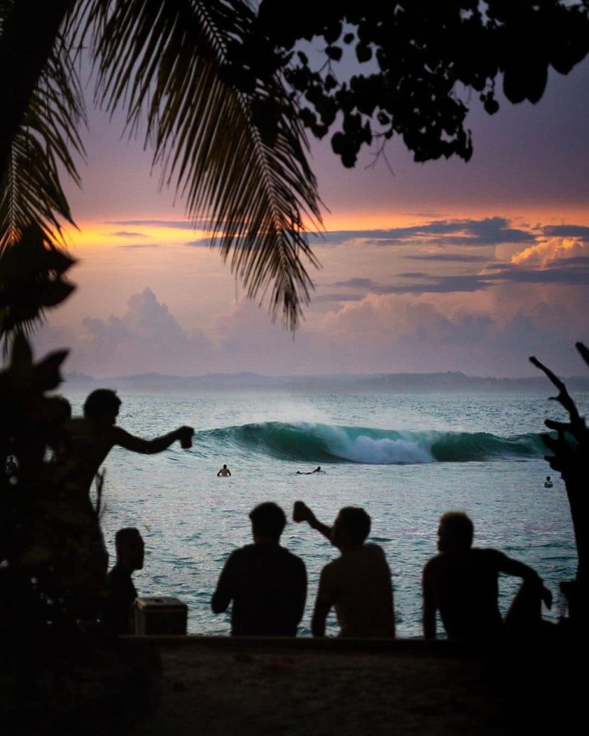 Friends hare a beer while watching surfers catch waves in fading sunlight after sunset.