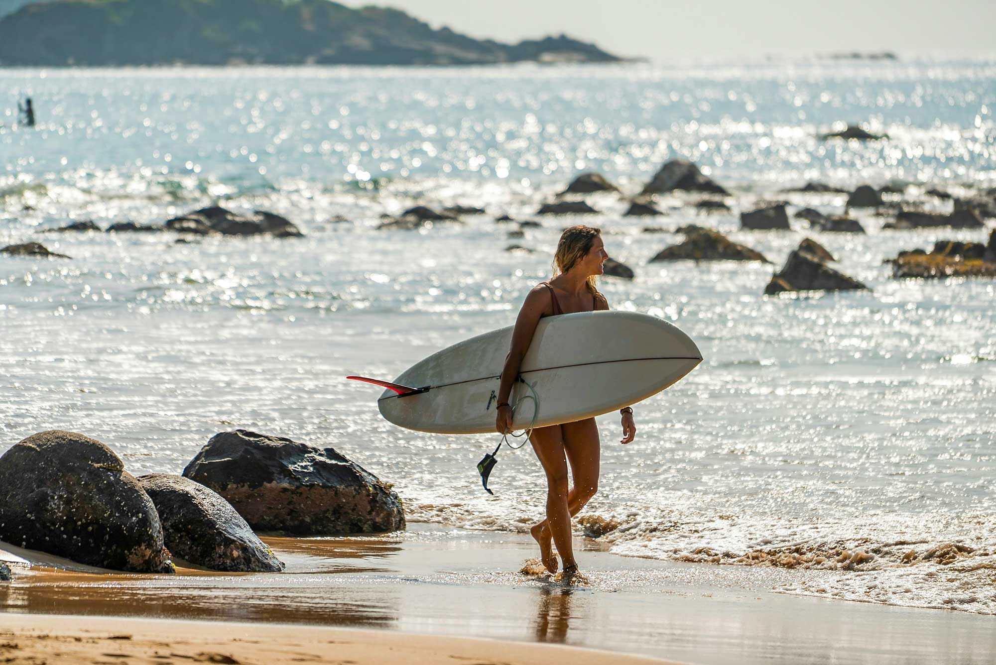 A girls walks along Waligama beach carrying a large white surfboard.